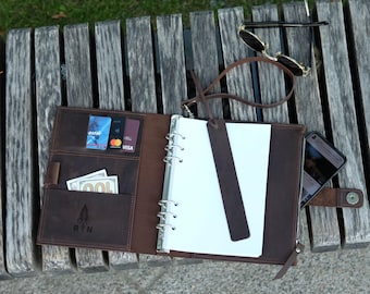 May include: A dark brown leather binder featuring a six-ring metal mechanism, multiple interior card slots, a cash pocket, and a matching leather bookmark. The cover displays a small embossed logo with the letters R and N above a leaf icon.