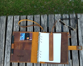 May include: A brown leather binder with a six-ring metal mechanism, a wrist strap, and a snap closure. The interior features card slots, a pen loop with a black pen, and a matching leather bookmark. The name Oscar is embossed on the cover.
