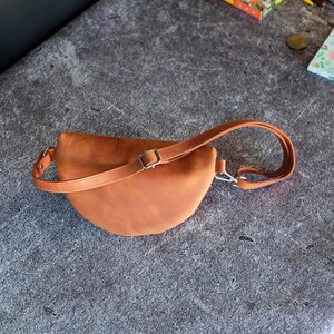 May include: A crescent-shaped tan leather fanny pack featuring a smooth matte finish, a single adjustable strap with a silver-toned metal buckle, and a silver-toned metal swivel clasp attachment on one side of the bag for secure closure.
