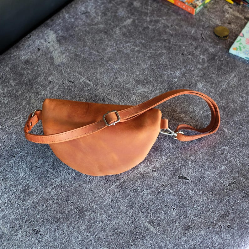 May include: A crescent-shaped tan leather fanny pack featuring a smooth matte finish, a single adjustable strap with a silver-toned metal buckle, and a silver-toned metal swivel clasp attachment on one side of the bag for secure closure.