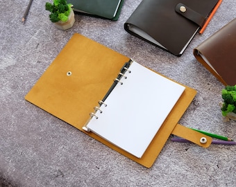 May include: An open mustard yellow leather binder with a six-ring metal mechanism holding blank white paper. The binder features a snap button closure strap. Additional closed leather binders in dark green and brown are visible nearby.