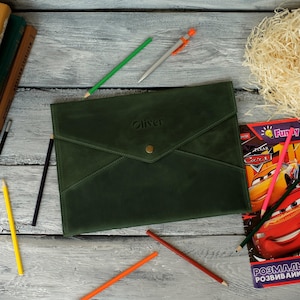 Könnte beinhalten: Eine dunkelgrüne Dokumentenmappe aus Leder in Umschlagform mit einer Klappe, einem messingfarbenen Druckknopf und dem eingeprägten Namen Oliver in der Mitte. Die Mappe ist von mehreren Buntstiften und einem Notizbuch umgeben.