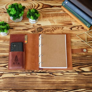 May include: A brown leather planner cover featuring a four-ring binder mechanism, a dedicated pocket containing a black smartphone, and a small card slot. The front pocket is embossed with a feather graphic and the letters R and N.