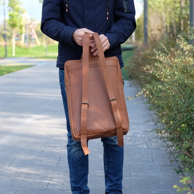 May include: A brown leather backpack with adjustable straps. The backpack has a flap closure and a zippered pocket on the front.