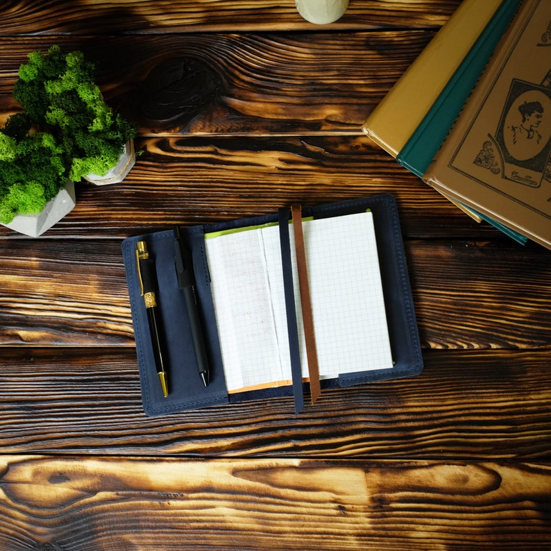 May include: A navy blue leather notebook with a grid-lined paper insert, open to display the pages. A gold and black pen and a black pen are inside the cover. A brown leather strap holds the notebook closed. The notebook is on a wooden surface.