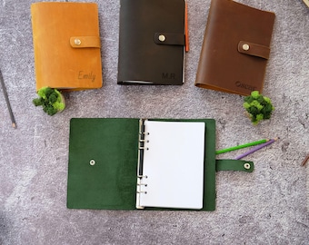 Puede incluir: Cuatro cuadernos de cuero con cierres de presión metálicos. Tres están cerrados y tienen nombres o iniciales: Emily en color canela, M.R. en marrón oscuro y Oscar en marrón medio. Un cuaderno verde está abierto, mostrando un mecanismo de seis anillas.