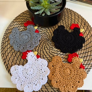 Peut inclure: Quatre dessous-de-verre en forme de poulet crochetés en gris, noir, blanc et marron. Les dessous-de-verre sont disposés sur un set de table en osier.