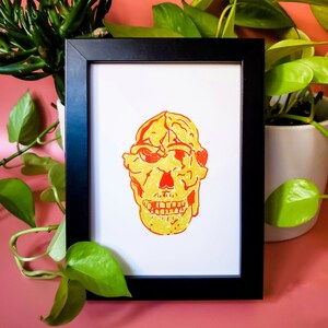 May include: A framed print of a yellow and red skull illustration. The skull is drawn in a simple style with a wide smile.