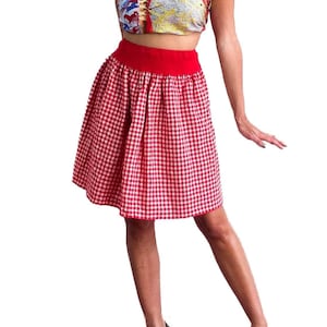 Peut inclure: Une femme portant une jupe à carreaux rouge et blanc avec une ceinture rouge, un haut sans bretelles avec un motif coloré et un collier de chaîne en or. Elle porte des talons noirs.