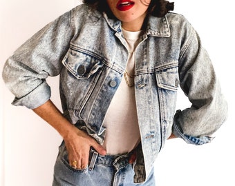May include: A light blue denim jacket with a faded, distressed look. The jacket has a button-up front, two chest pockets, and two front pockets. The model is wearing a white t-shirt underneath the jacket.