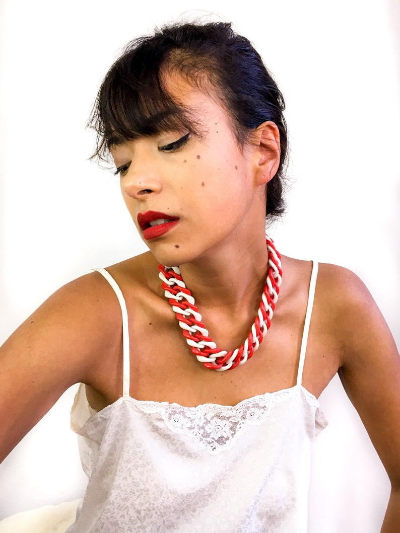 May include: A woman wearing a white tank top with a red and white plastic chain necklace. The necklace is thick and chunky.
