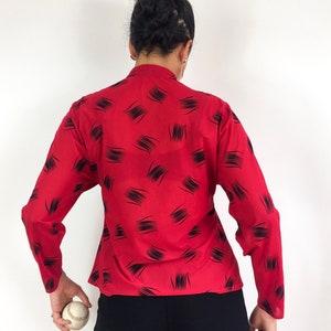 May include: Red long-sleeve blouse with a black abstract print. The blouse has a high neckline and a fitted silhouette.