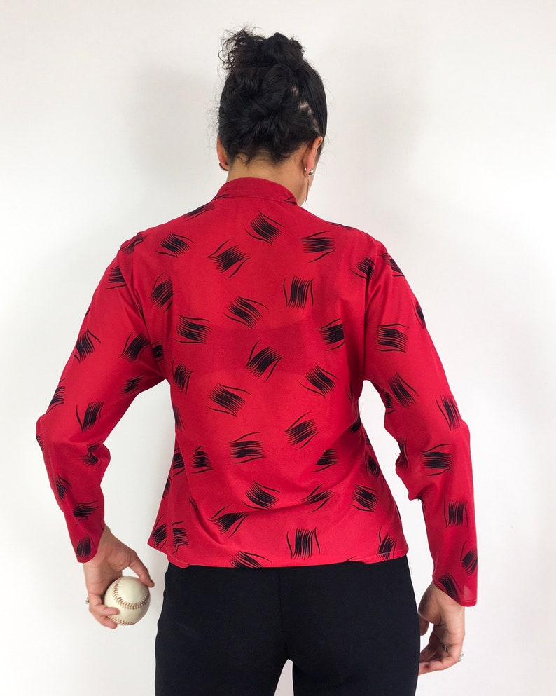 May include: Red long-sleeve blouse with a black abstract print. The blouse has a high neckline and a fitted silhouette.