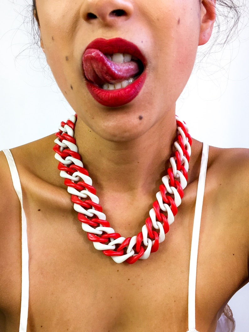 May include: A close-up of a person's mouth with red lipstick and a tongue sticking out. The person is wearing a red and white chunky chain necklace.