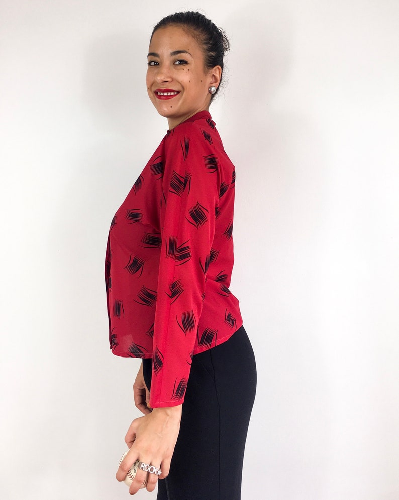 May include: A red long-sleeved blouse with a black abstract print. The blouse has a high neckline and a button-up front.