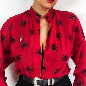 May include: A red button-down shirt with a black abstract print. The shirt is unbuttoned and has a collar. The person wearing the shirt is also wearing a black belt with a silver buckle.