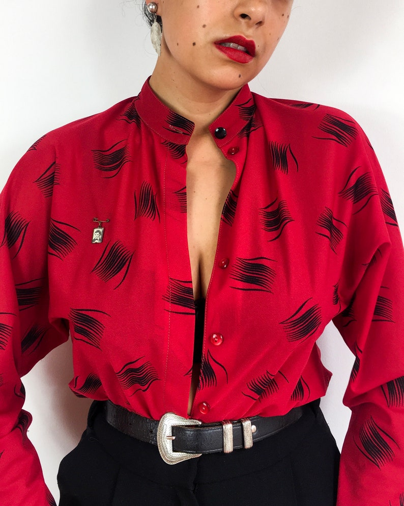 May include: A red button-down shirt with a black abstract print. The shirt is unbuttoned and has a collar. The person wearing the shirt is also wearing a black belt with a silver buckle.
