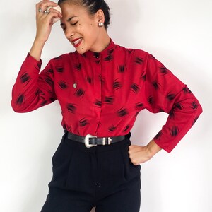 May include: A red long-sleeved blouse with a black abstract print and a black belt. The blouse has a high collar and a button-up front.