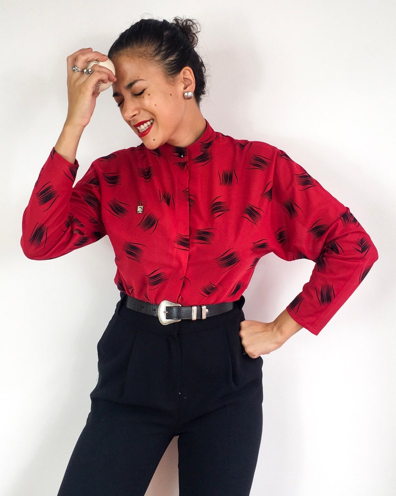 May include: A red long-sleeved blouse with a black abstract print and a black belt. The blouse has a high collar and a button-up front.