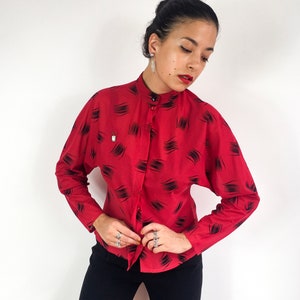 May include: A red long-sleeved button-up shirt with a black abstract print. The shirt has a mandarin collar and a fitted silhouette.