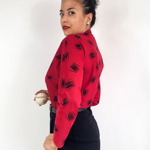 May include: A woman wearing a red long-sleeved blouse with a black abstract print and black pants. She is holding a white baseball.