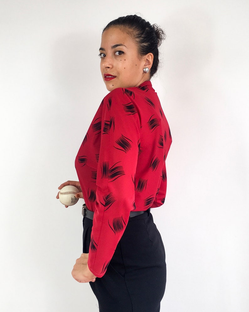 May include: A woman wearing a red long-sleeved blouse with a black abstract print and black pants. She is holding a white baseball.