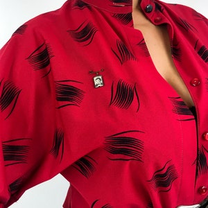 May include: A red button-up shirt with a black abstract pattern. The shirt has a high collar and a long sleeve. A small gold pin with a picture of a person is on the shirt.