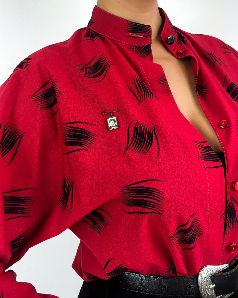 May include: A red button-up shirt with a black abstract pattern. The shirt has a high collar and a long sleeve. A small gold pin with a picture of a person is on the shirt.