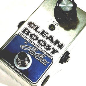 May include: A silver metal guitar effects pedal labeled CLEAN BOOST, HANDWIRED IN DEVON, ENGLAND, and Beller Instruments. It features a single black control knob, a small active indicator light, a metallic footswitch, and a blue graphic panel.