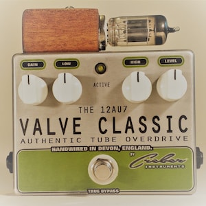 May include: A silver and green guitar pedal with the text "THE 12AU7 VALVE CLASSIC AUTHENTIC TUBE OVERDRIVE HANDWIRED IN DEVON, ENGLAND. BY Creeber INSTRUMENTS TRUE BYPASS" on the front. The pedal has four knobs labeled "GAIN", "LOW", "HIGH", and "LEVEL".