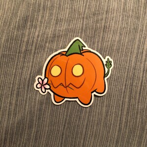 May include: A cartoon sticker of an orange pumpkin with a green stem and a smiling face. The pumpkin has two yellow eyes and is holding a pink flower in its mouth. The sticker has a white border.