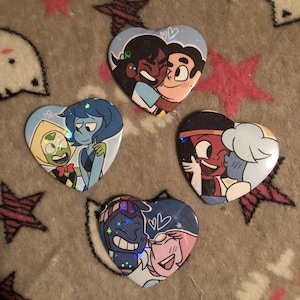 May include: Four heart-shaped buttons featuring cartoon characters in various embraces. Each button has a blue background with glitter accents. The characters are from the animated series Steven Universe. The buttons are on a gray patterned surface.