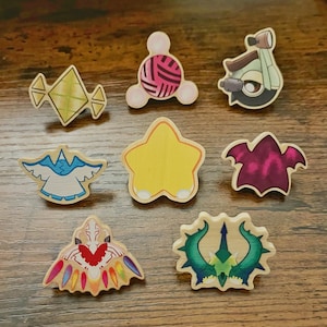 May include: A collection of wooden pins with various designs. The pins include a yellow star, a pink ball of yarn, a green and yellow diamond, a blue and white bird, a purple bat, a green trident, a brown scooter, and a colorful winged creature.