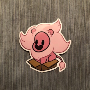 May include: A sticker featuring a cartoon lion with a pink mane and body, sitting inside a brown cardboard box. The lion has a black smiling mouth and eyes. The sticker is on a blue fabric background.
