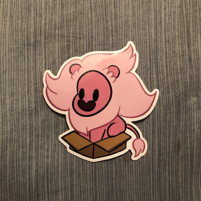May include: A sticker featuring a cartoon lion with a pink mane and body, sitting inside a brown cardboard box. The lion has a black smiling mouth and eyes. The sticker is on a blue fabric background.