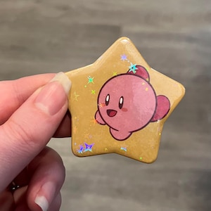 May include: A star-shaped pin featuring a pink, round cartoon character with large eyes and a smiling mouth. The background of the pin is a golden yellow color with a holographic star pattern that reflects light, creating small rainbow-colored sparkles.