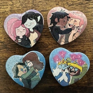 May include: Four heart-shaped badges with cartoon couples. Each badge depicts a different pair, with diverse skin tones and hair colours. The backgrounds are shades of pink and blue, with small hearts and stars.