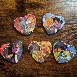 May include: Five heart-shaped pinback buttons featuring colorful cartoon illustrations of characters in pairs. Each button has a holographic star finish. The designs depict various animated figures embracing or interacting, with small red heart icons.