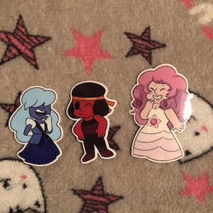 May include: Three cartoon character stickers. One is blue with light blue hair, one is red with black hair, and the last is pink with curly pink hair. The stickers are on a grey background with pink star shapes.