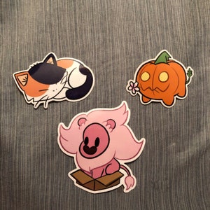 May include: Three cartoon stickers: a sleeping calico cat, an orange jack-o'-lantern with a flower, and a pink lion in a box. The stickers are on a blue-gray fabric background.