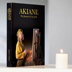 May include: A black book titled "AKIANE The Journey of an Artist" with gold lettering. The cover features a painting of a woman in a yellow outfit holding a paintbrush and palette. A lit white candle is to the right.