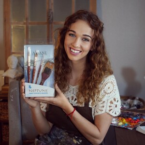 May include: A woman holds a box of Neptune paint brushes. The box is white with blue accents and features the brand name "Neptune" and the text "Artist Paint Brushes." The brushes are made of natural hair and have wooden handles.