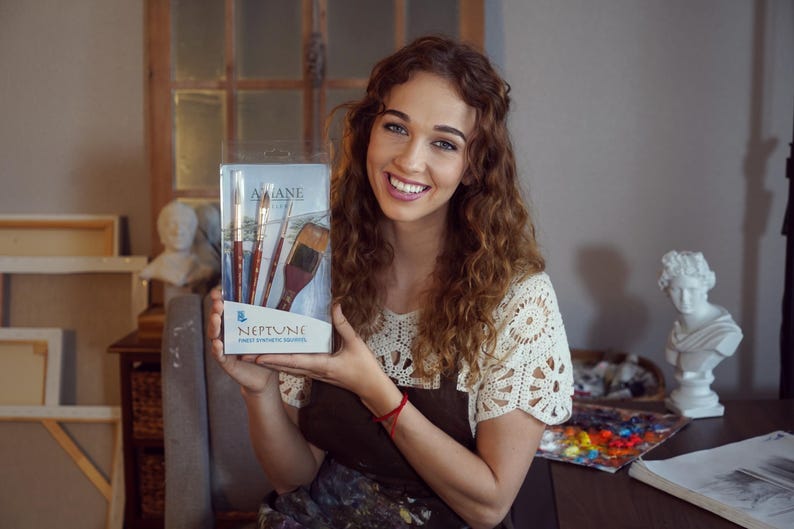 May include: A woman holds a box of Neptune paint brushes. The box is white with blue accents and features the brand name "Neptune" and the text "Artist Paint Brushes." The brushes are made of natural hair and have wooden handles.