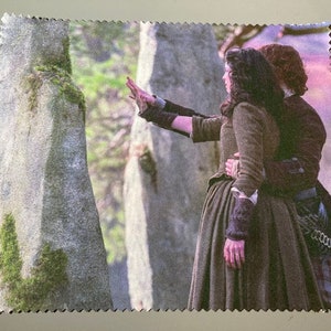 May include: A couple stand in front of a stone structure, the woman in a brown dress and the man in a kilt. The woman is reaching out with her left hand, while the man has his arm around her waist.