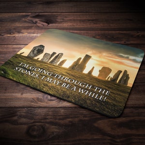 May include: A brown and black mouse mat with a picture of a stone circle at sunset. The text "I'm going through the stones, I may be a while!" is printed on the mouse mat.