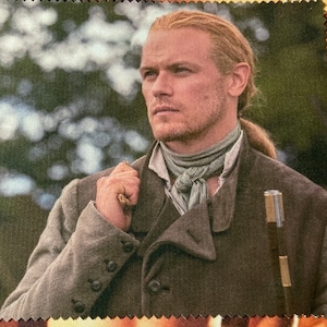 Op de afbeelding: Een man met lang, rood haar draagt een bruine jas met een witte sjaal. Hij houdt een zilveren en bruine wandelstok vast.