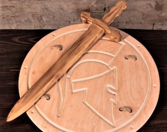 May include: A wooden toy sword and shield set. The shield is round and features a carved Spartan helmet design. The sword is made of wood and has a simple design.