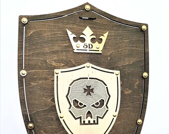 May include: A wooden shield with a gold skull and crossbones design in the centre. The shield is decorated with gold studs and a gold crown with the letters "SD" on top.