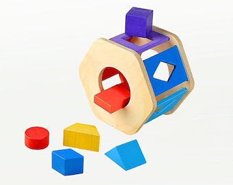Op de afbeelding: Een houten vormsorteerspeelgoed met een natuurlijke houten afwerking. Het speelgoed heeft verschillende gekleurde geometrische vormen, waaronder een rode cilinder, een geel huis, een blauwe kubus en een blauwe driehoek. De sorteerder heeft openingen voor elke vorm.