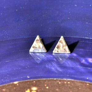 May include: A pair of silver triangle stud earrings. The earrings have a textured surface and are set against a dark blue background. The earrings are small and geometric.